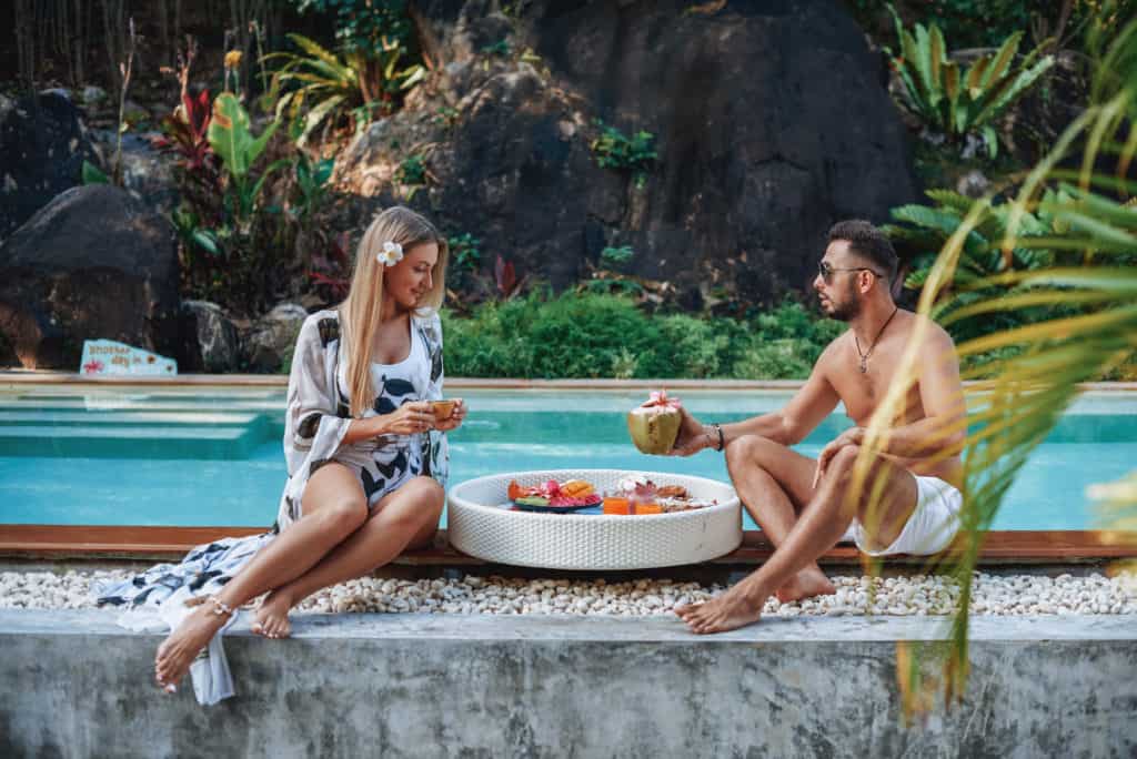 Relaxed couple of tourists in luxurious hotel eat asian food and drink juice in background of jungle and swimming pool.